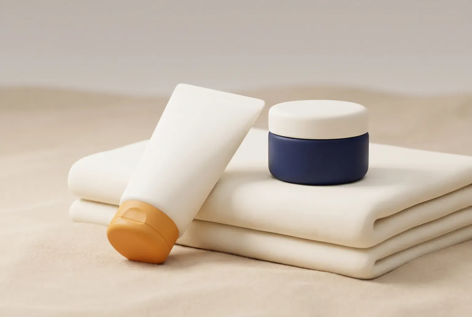Illustration: a plain cream squeeze tube with an amber cap and a navy lotion jar on a folded cream towel on soft sand, drawn as a soft 3D render. Not a real product.