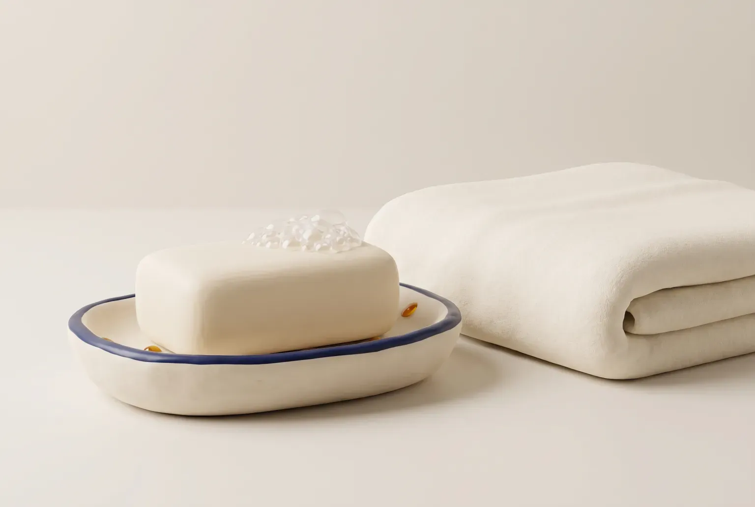 Illustration: a plain white rectangular bar of soap with soft foam bubbles on a simple ceramic soap dish beside a folded cream towel, drawn as a soft 3D render. Not a real product.
