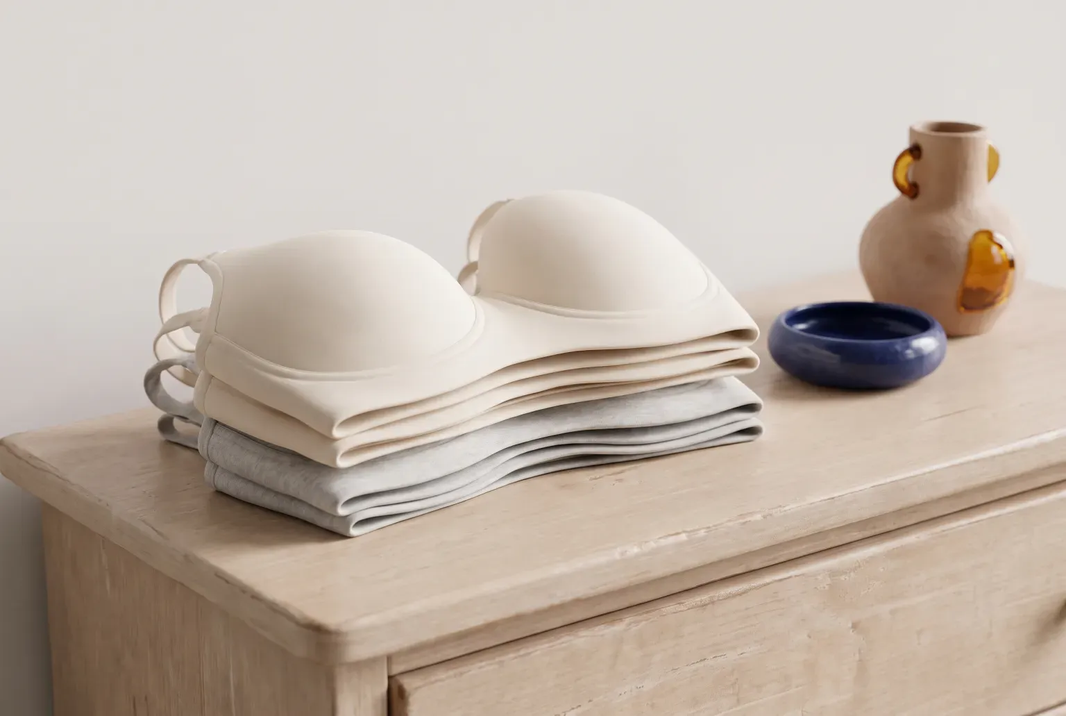 Illustration: a stack of folded cream and grey soft bras on a pale wooden dresser beside a navy bowl and a small vase, drawn as a soft 3D render. Not a real product.