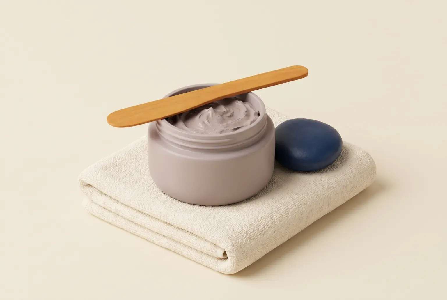 Illustration: a small round jar of soft grey-pink clay mask with a wooden spatula resting across it, on a folded cream towel beside a smooth navy stone, drawn as a soft 3D render. Not a real product.