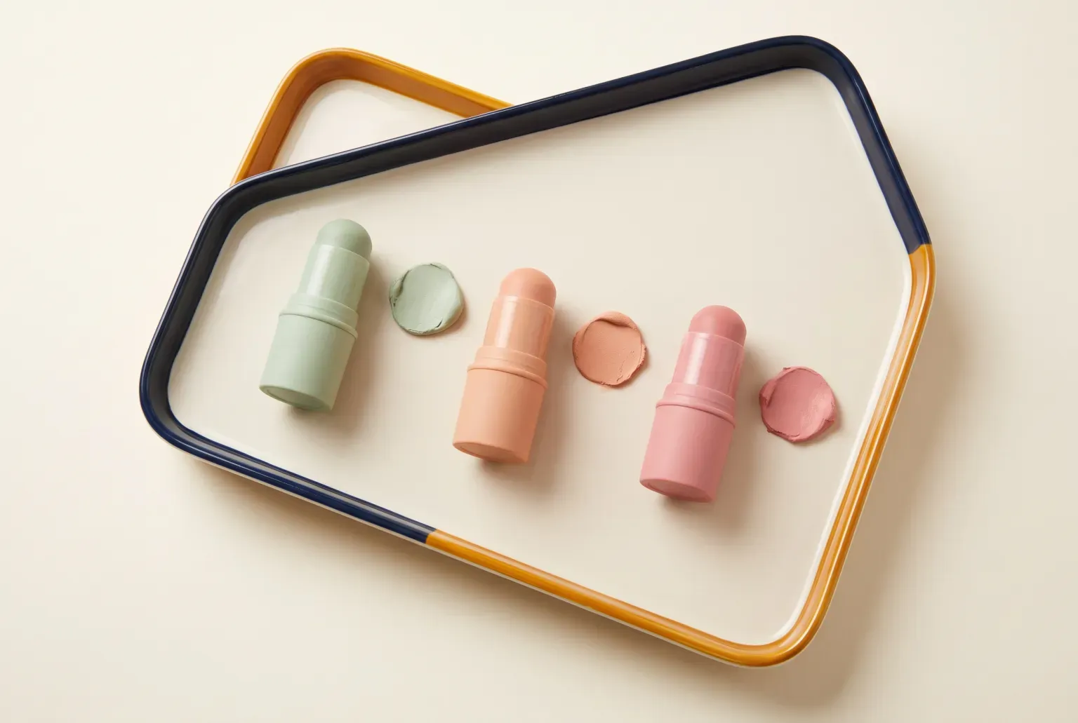Illustration: three pastel makeup sticks in green, peach and pink with a matching swatch beside each, laid on a cream and navy tray, drawn as a soft 3D render. Not a real product.