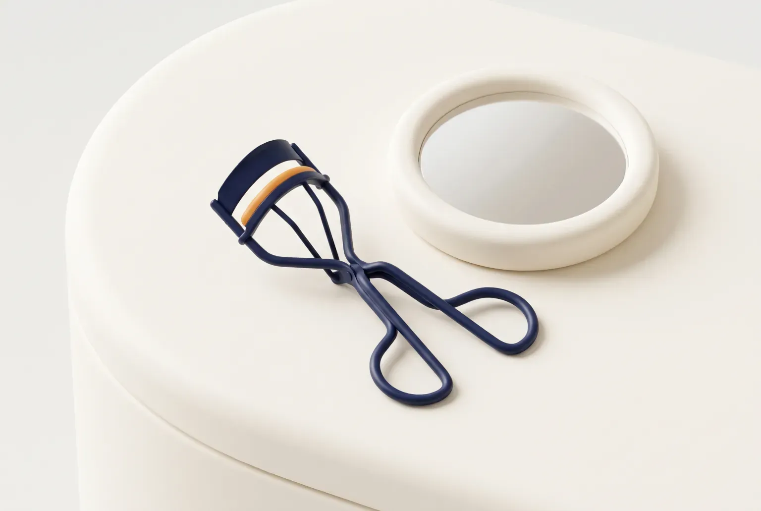 Illustration: a plain metal eyelash curler with a soft rubber pad lying beside a small mirror on a vanity, drawn as a soft 3D render. Not a real product.
