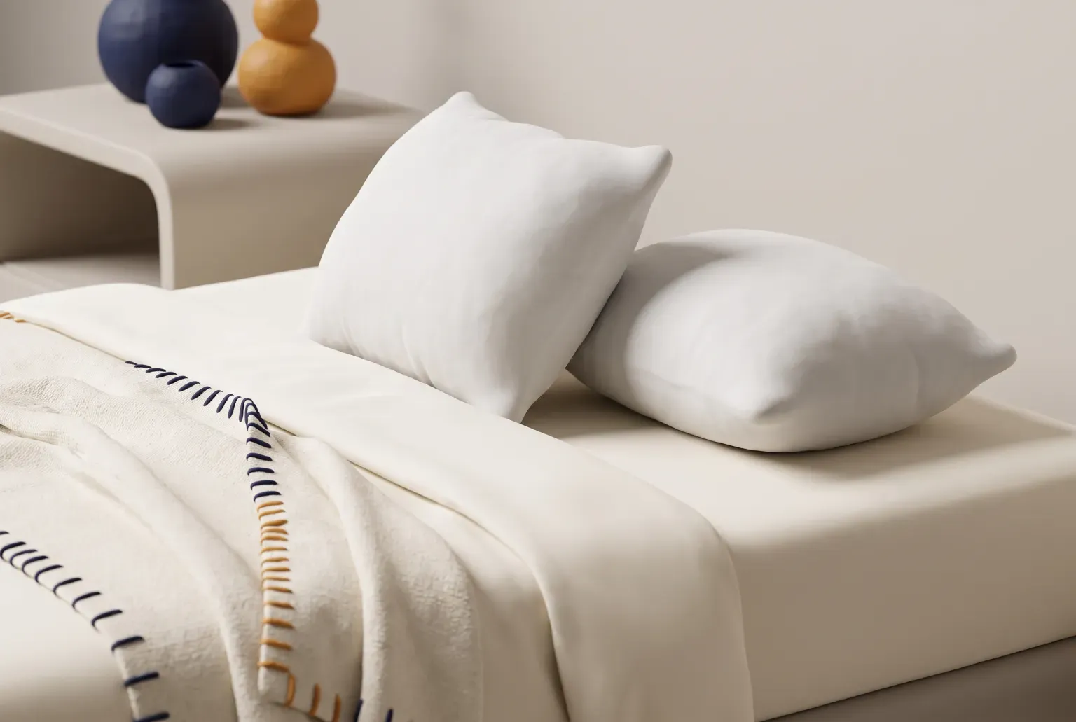 Illustration: two plain white pillows on a made bed beside a cream blanket with navy and orange stitching, drawn as a soft 3D render. Not a real product.