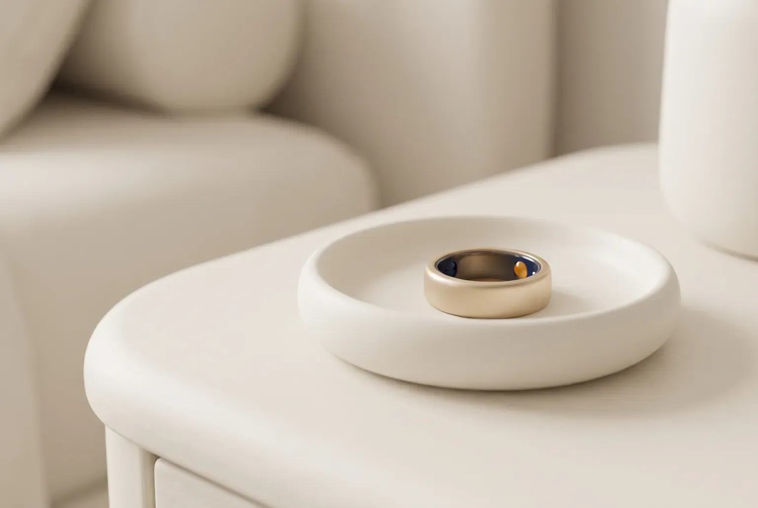 Illustration: a slim smooth smart ring in a soft gold tone resting on a small round cream dish on a bedside table beside a pale armchair, drawn as a soft 3D render. Not a real product.